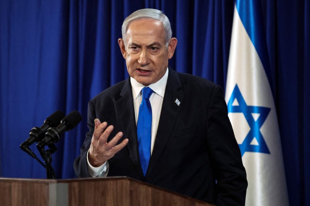Man in a dark suit and bright blue tie speaks at a podium with microphones blue curtains and an Israeli flag in the background