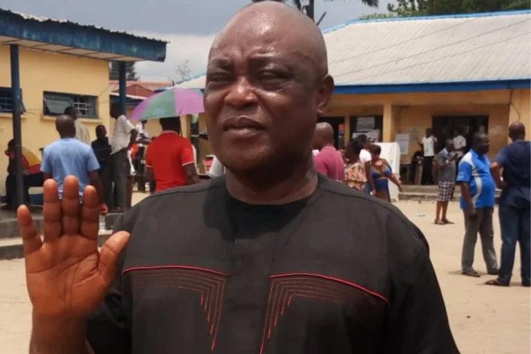 Man in a dark shirt faces the camera with his hand raised, in a busy outdoor market setting behind him.