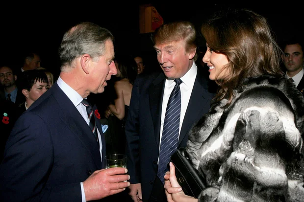 Three formally dressed people chat at a nighttime reception a man on the left holds a glass the middle man speaks and a woman in a fur coat smiles nearby