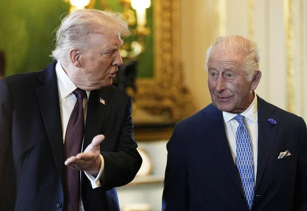 Two well-dressed men in a formal room engaged in conversation; the left man gestures with his hand.